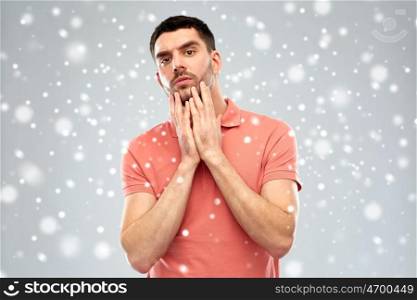 facial expression, winter, christmas and people concept - young man in polo t-shirt touching his face over snow on gray background