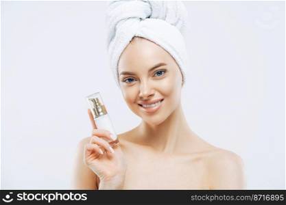 Facial treatment, cosmetology, beauty concept. Beautiful smiling woman poses with cosmetic product indoor, has healthy well cared skin, wears wrapped towel on head, enjoys rejuvenation treatment