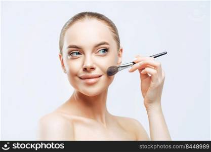 Facial treatment, cosmetology concept. Pretty young woman with dark combed hair, applies powder foundation on face with beauty brush, stands sideways, has naked body, isolated on white background
