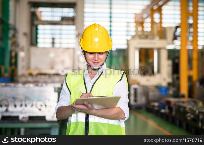 Factory technician famale inspector write audit report on on clipboard for internal examine with blur warehouse machinery background. Quality assurance (QA) for safety business process.