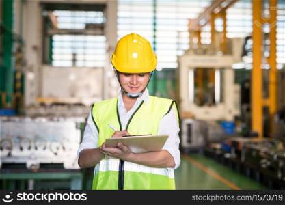 Factory technician famale inspector write audit report on on clipboard for internal examine with blur warehouse machinery background. Quality assurance (QA) for safety business process.