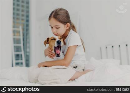 Fair haired small girl with pony tail wears casual pyjamas, embraces dog who yawns, pose together on comfortable bed, expresses love and care to domestic animal, spends morning weekend at home