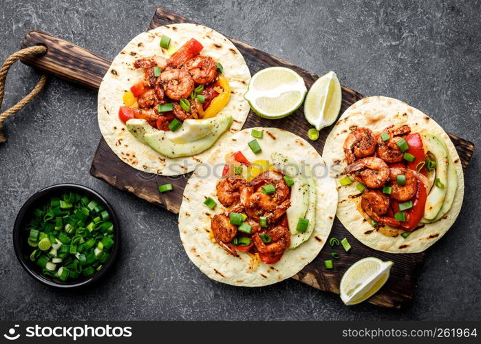 Fajitas in tortillas with fried shrimps, bell peppers and onion served up with avocado and green onions on wooden cutting board, top view, flat lay