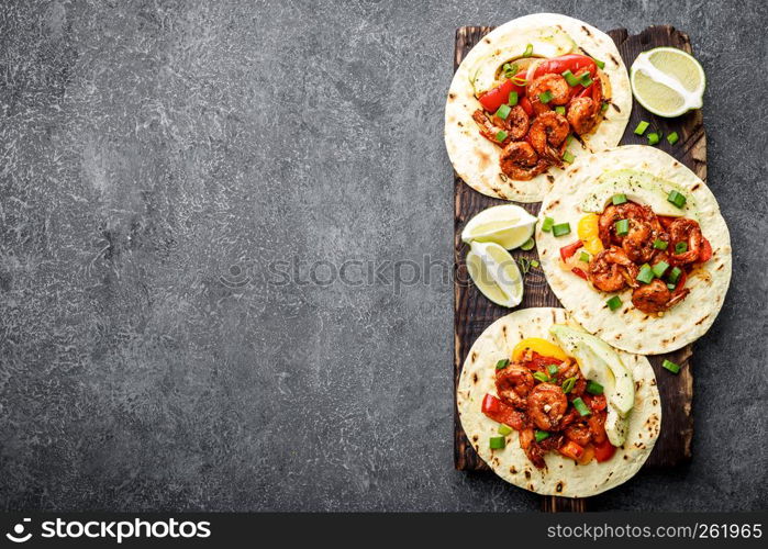 Fajitas in tortillas with fried shrimps, bell peppers and onion served up with avocado and green onions on wooden cutting board, top view, food background with space for a text