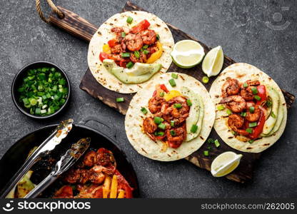 Fajitas in tortillas with fried shrimps, bell peppers and onion served up with avocado and green onions on wooden cutting board, top view, flat lay