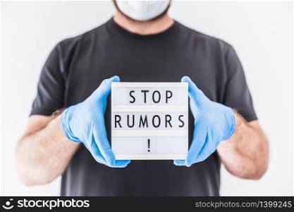 Fake news infodemics during Covid-19 pandemic concept. Man wearing protective mask and medical gloves on hands holding lightbox with text Stop rumors. People want to know the truth about coronavirus