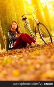 Fall active lifestyle concept,. Beauty young redhaired woman fashion girl relaxing in autumn park with bicycle, sitting alone under tree, outdoor