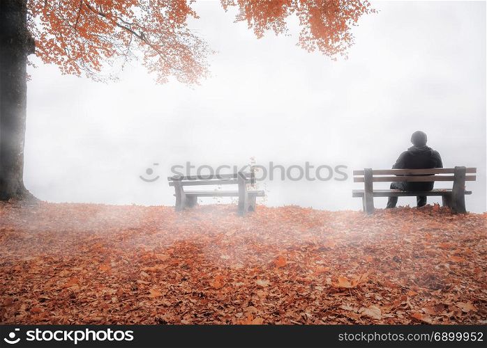 Fall image with a man sitting alone, on a wooden bench, thinking, shrouded by a dense mist and surrounded by autumn colorful nature.
