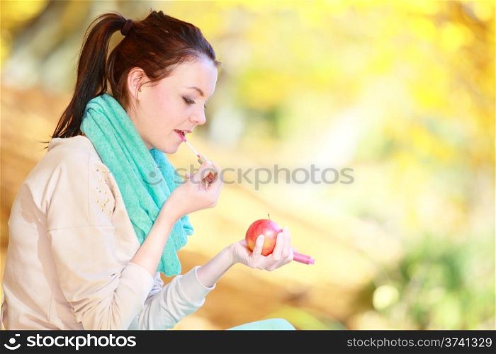 Fall lifestyle concept, harmony freedom. Casual young woman girl relaxing in autumnal park applying lipstick holding red apple. Golden colorful leaves background
