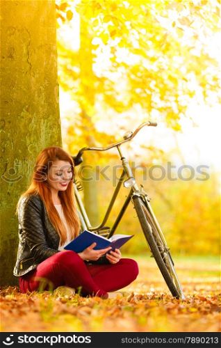 Fall lifestyle concept. Redhaired young woman girl relaxing in autumn park reading book, sitting alone under tree. Sunny day