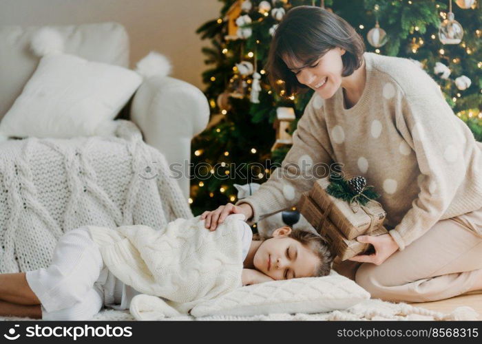 Famaily, happy winter holidays concept. Caring mom stands on knees near sleeping daughter, prepares presents for New Year, has pleasant smile, pose in cozy living room. Christmas night and miracle