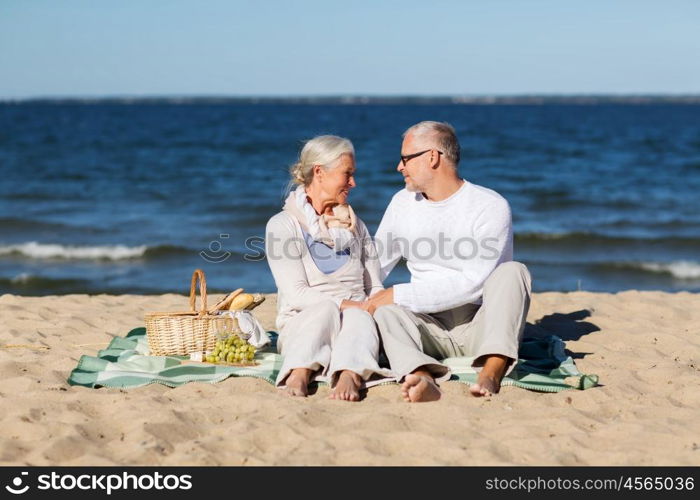 family, age, holidays, leisure and people concept - happy senior couple with picnic basket sitting on blanket on summer beach