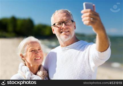 family, age, travel, technology and people concept - happy senior couple with smartphone taking selfie and hugging on summer beach