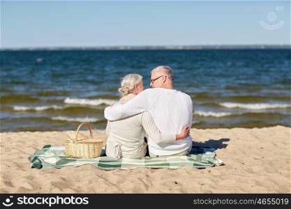 family, age, travel, tourism and people concept - happy senior couple sitting on plaid with picnic basket and hugging on summer beach