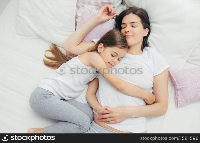 Family and bed time concept. Pleased sleepy mother and her adorable daughter embrace in bed and have pleasant dreams, lie on white bedclothes, have good relationships, take care of each other