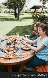 Family and friends having meal - pizza, salads, fruits and drinking white wine during summer picnic outdoor dinner in a home garden. Close up of people sitting at the table in a orchard in a backyard. Family and friends having meal - pizza, salads, fruits and drinking white wine during summer picnic outdoor dinner in a home garden