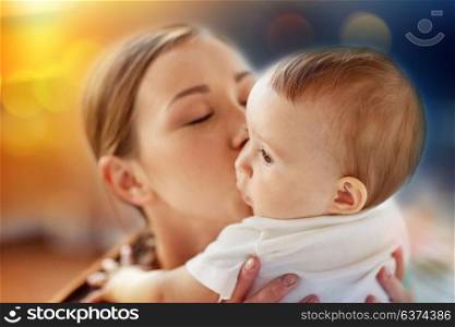 family and motherhood concept - close up of happy young mother kissing little baby over lights background. close up of happy young mother kissing little baby