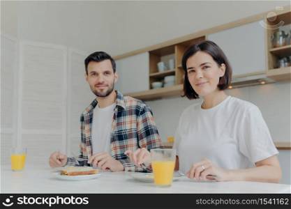 Family and nutrition concept. Husband and wife look joyfully at camera, enjoy delicious breakfast, drink fresh made juice, discuss their family plans, pose in modern kitchen, cozy atmosphere