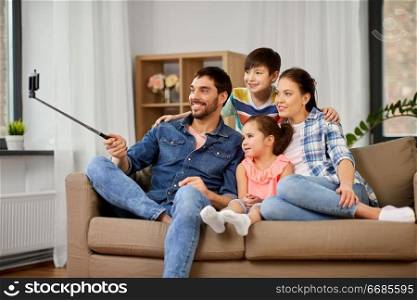 family and people concept - happy father, mother, little son and daughter taking picture by smartphone on selfie stick at home. happy family taking selfie at home