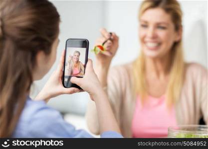 family and people concept - happy girl with smartphone having dinner and photographing her mother at home kitchen. girl photographing mother by smartphone at home. girl photographing mother by smartphone at home