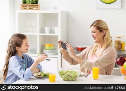 family and people concept - happy mother with smartphone having dinner and photographing her daughter at home kitchen. woman photographing daughter by smartphone at home