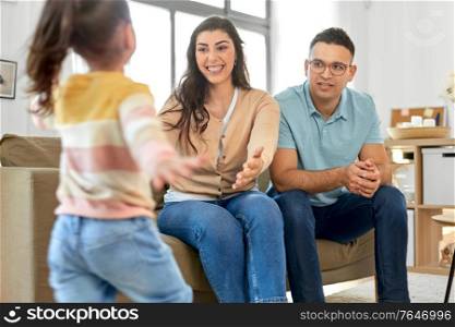 family and people concept - little daughter running to happy smiling mother and father sitting on sofa at home. happy family at home