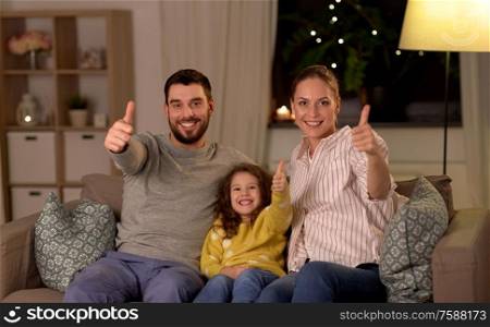 family and people concept - portrait of happy smiling father, mother and little daughter sitting on sofa and showing thumbs up at home at night. portrait of happy family sitting on sofa at home
