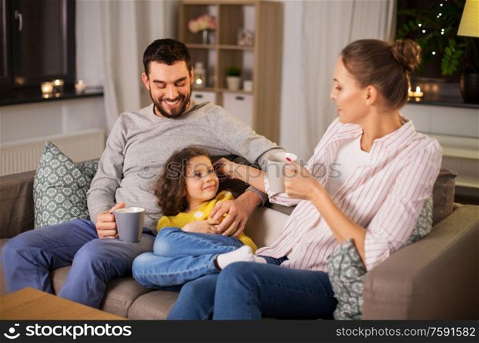 family and people concept - portrait of happy smiling father, mother drinking tea and little daughter sitting on sofa at home at night. portrait of happy family sitting on sofa at home