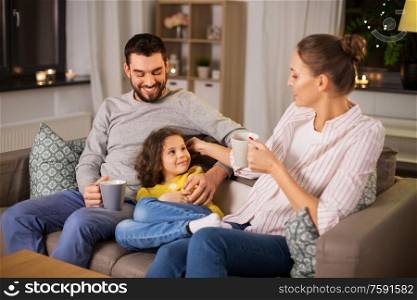 family and people concept - portrait of happy smiling father, mother drinking tea and little daughter sitting on sofa at home at night. portrait of happy family sitting on sofa at home