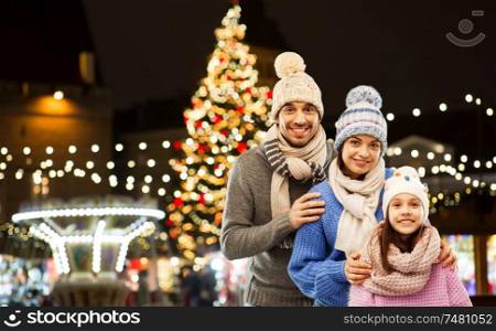 family and winter holidays concept - happy mother, father and little daughter in knitted hats and scarves over christmas market lights in old town of tallinn city background. happy family over christmas market in tallinn