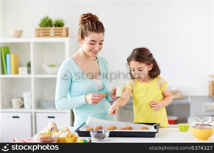 family, baking, cooking and people concept - happy mother and little daughter eating cookies with milk at home kitchen. happy mother and daughter eating cookies at home