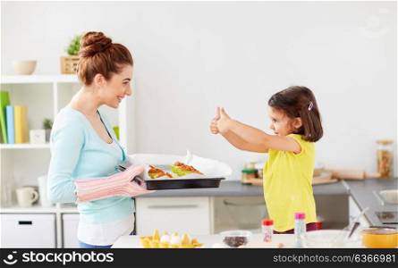 family, baking, cooking and people concept - mother holding tray of muffins and little daughter showing thumbs up at home kitchen. mother and daughter baking muffins at home