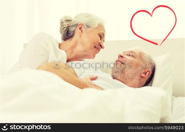 family, bedtime, valentines day, love and people concept - happy senior coupler lying in bad and talking at home with red heart shape
