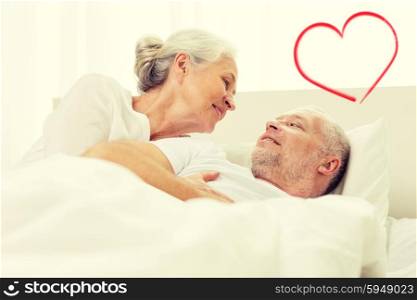 family, bedtime, valentines day, love and people concept - happy senior coupler lying in bad and talking at home with red heart shape