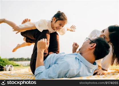 Family bonds over laughter and joy in the park. Father lifts his daughter high, as she happily flies like a plane with arms spread wide, and mother watches with pride. A picture of family fun and love