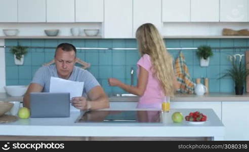 Family budget, bills and finances. Stressed man with laptop computer analyzing bills at home while holding his head with hands and looking at documents. Wife and daughter on bckground. Father reviewing family expenses using laptop at modern kitchen.