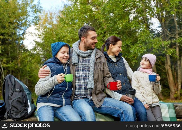family, camping and tourism concept - happy mother, father, son and daughter sitting on bench and drinking hot tea from cups at camp in woods. happy family with cups drinking hot tea at camp