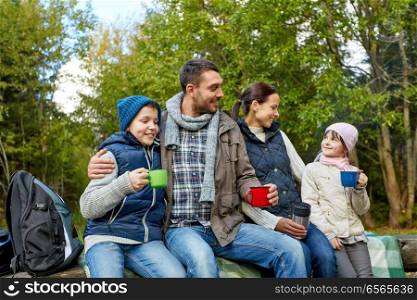 family, camping and tourism concept - happy mother, father, son and daughter sitting on bench and drinking hot tea from cups at camp in woods. happy family with cups drinking hot tea at camp