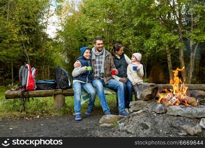 family, camping and tourism concept - happy mother, father, son and daughter sitting on bench and drinking hot tea from cups at camp fire in woods. happy family drinking hot tea near camp fire. happy family drinking hot tea near camp fire