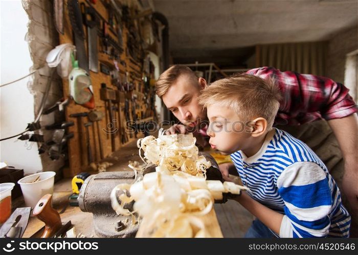 family, carpentry, woodwork and people concept - father and little son blowing shavings off wood plank at workshop