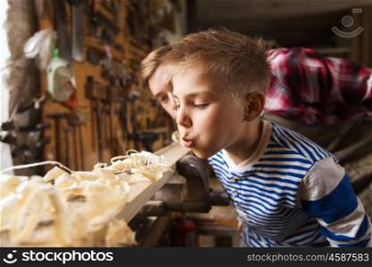 family, carpentry, woodwork and people concept - father and little son blowing shavings off wood plank at workshop