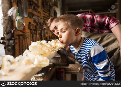 family, carpentry, woodwork and people concept - father and little son blowing shavings off wood plank at workshop