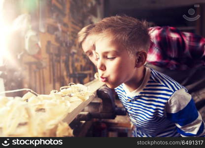 family, carpentry, woodwork and people concept - father and little son blowing shavings off wood plank at workshop. father and little son with wood plank at workshop. father and little son with wood plank at workshop