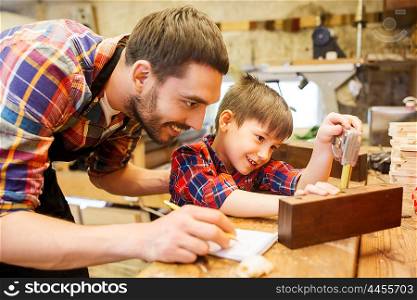 family, carpentry, woodwork and people concept - father and little son with ruler and pencil measuring wood plank and writing dimensions to notebook at workshop