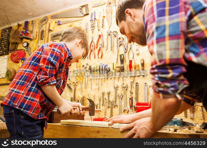 family, carpentry, woodwork and people concept - father and little son with hammer hammering nail into wood plank at workshop