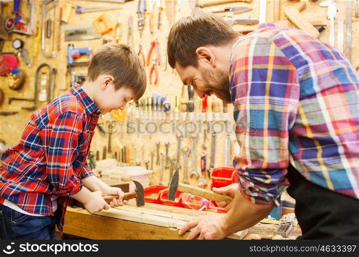 family, carpentry, woodwork and people concept - father and little son with hammer hammering nail into wood plank at workshop