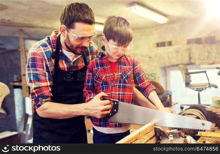 family, carpentry, woodwork and people concept - father and little son with saw sawing wood plank at workshop. father and son with saw working at workshop. father and son with saw working at workshop