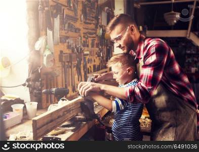 family, carpentry, woodwork and people concept - father and little son with plane working with wood plank at workshop. father and son with plane shaving wood at workshop. father and son with plane shaving wood at workshop
