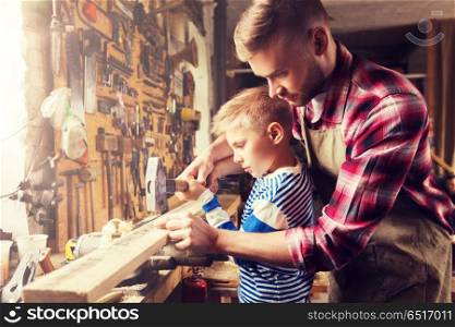 family, carpentry, woodwork and people concept - father and little son with hammer hammering nail into wood plank at workshop. father and son with hammer working at workshop. father and son with hammer working at workshop