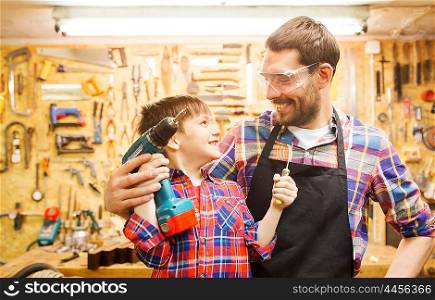 family, carpentry, woodwork and people concept - happy father and little son with drill and screwdriver working at workshop